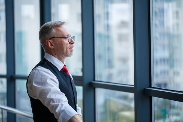 Улыбаясь, счастливый управляющий директор думает о своем успешном карьерном развитии, стоя в своем офисе на фоне окна с копировальным пространством
 - Фото, изображение