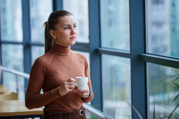Mosolygó boldog ügyvezető nő igazgató gondolkodás a sikeres karrierjét, miközben áll egy csésze aromás kávé az irodájában, közel a háttérben egy ablak másolási hely - Fotó, kép
