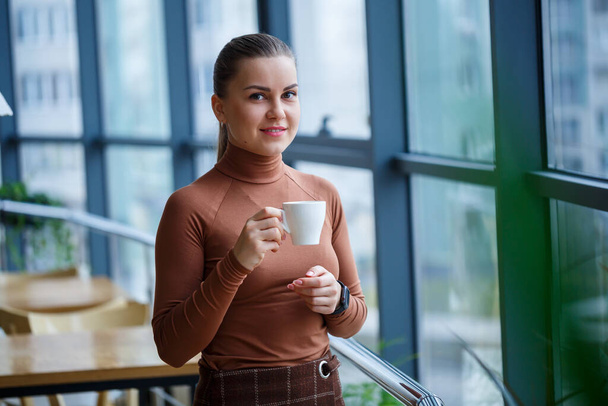 Lächelnd denkt die Geschäftsführerin an ihre erfolgreiche berufliche Entwicklung, während sie mit einer Tasse aromatischem Kaffee in ihrem Büro vor einem Fenster mit Kopierraum steht. - Foto, Bild