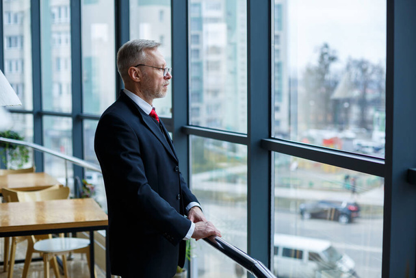 Adult male businessman, teacher, mentor looks out the large window of his office. Work in the office. Development of a new business project - Photo, Image