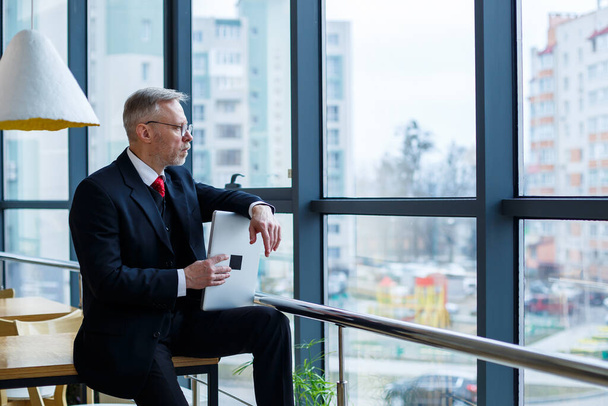 Улыбаясь счастливый управляющий директор думает о своем успешном развитии карьеры, стоя с ноутбуком в своем офисе на фоне окна с копировальным местом
 - Фото, изображение
