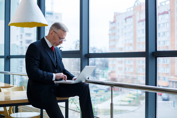 Улыбаясь счастливый управляющий директор думает о своем успешном развитии карьеры, стоя с ноутбуком в своем офисе на фоне окна с копировальным местом
 - Фото, изображение