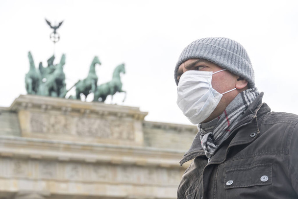 Ausbruch der Krankheit, Coronavirus covid-19 Pandemie. Porträt eines erwachsenen Mannes mit medizinischer Schutzmaske im Gesicht und dem Brandenburger Tor im Hintergrund - Foto, Bild