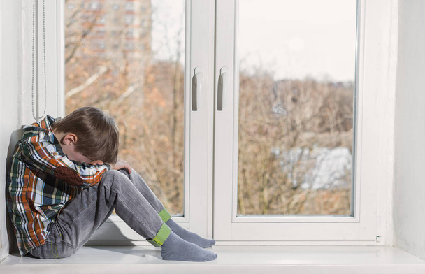 Sad young boy sitting in window and hide his face in hands. - Foto, afbeelding