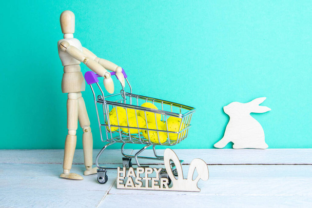 Het Paasconcept. Figuur van een houten man met een mand gevuld met eieren. Op de achtergrond is een houten konijn en een teken van een Gelukkig Pasen. - Foto, afbeelding