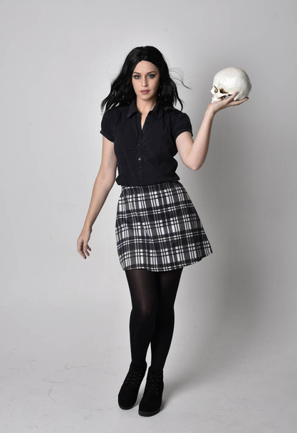 Πορτρέτο ενός goth κοριτσιού με σκούρα μαλλιά φορώντας μπλε και καρό φούστα με μπότες. Στέκεται όρθιος, κρατώντας ένα ανθρώπινο κρανίο, σε φόντο στούντιο. - Φωτογραφία, εικόνα