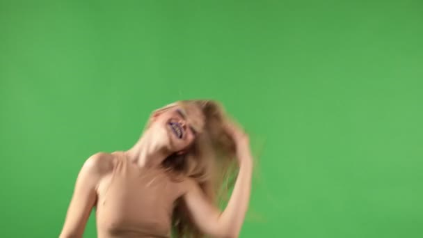 Krásná mladá žena ve stylovém oblečení potřásá hlavou s dlouhými blond vlasy a tancuje s úsměvem na kameru a izolované zelené pozadí - Záběry, video