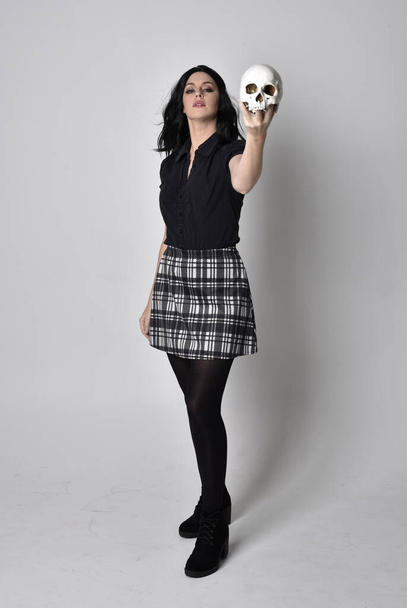 Портрет готической девушки с темными волосами в голубой клетчатой юбке с сапогами. Полностью стоящая поза, держащая человеческий череп, на фоне студии
. - Фото, изображение