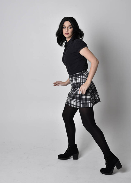 Portrait of a goth girl with dark hair wearing blue and plaid skirt with boots. Full length standing pose on a studio background. - Foto, Imagen