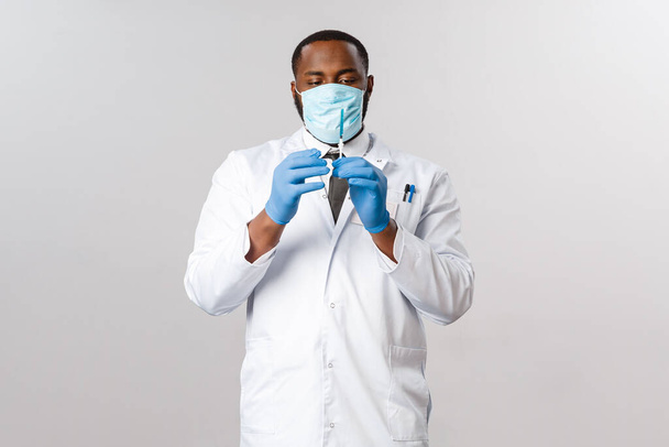 Covid-19, tratamiento del paciente con coronavirus y concepto de laboratorio. Retrato del guapo médico afroamericano profesional en mascarilla médica y guantes de látex, sostener la jeringa con la vacuna
 - Foto, imagen