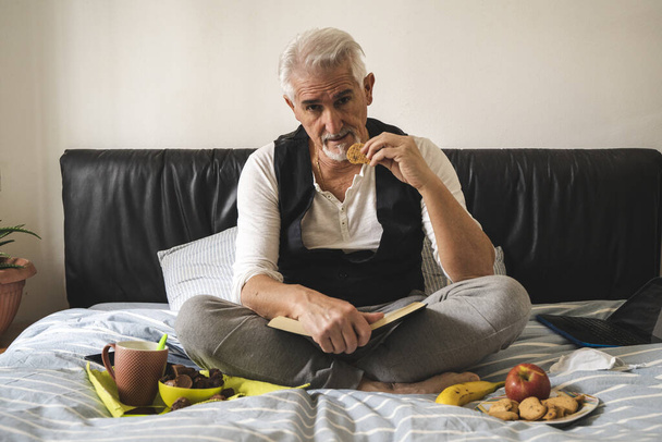 mature man is relaxing having breakfast in the bed of his home during tough quarantine at the time of coronavirus - Fotografie, Obrázek