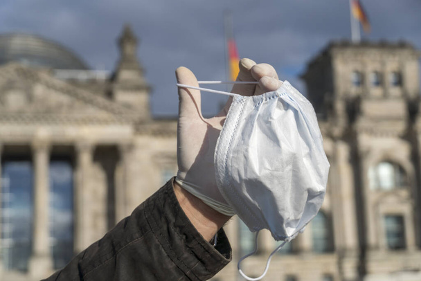 Manos de hombre cubiertas por un guante de látex que muestra una máscara de protección médica fuera del Bundestag alemán en Berlín. Enfoque selectivo
 - Foto, Imagen