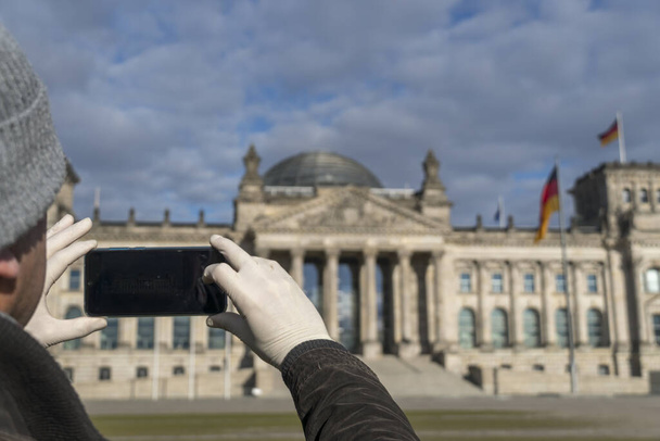 Man's hands covered by medical protective gloves taking a picture of the Berlin Bundestag building with his smartphone - Photo, Image