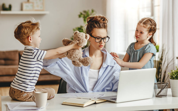 Upset girl hugging and small son showing toy to busy mother freelancer sitting at table with cup of coffee and using laptop against blurred interior of light modern apartmen - Фото, изображение