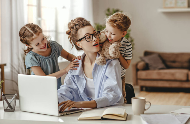 Upset girl hugging and small son showing toy to busy mother freelancer sitting at table with cup of coffee and using laptop against blurred interior of light modern apartmen - Foto, afbeelding