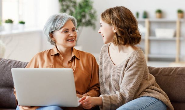 Happy aged woman with young adult daughter smiling and looking at each other while sitting on sofa and using laptop in living roo - Foto, Imagen