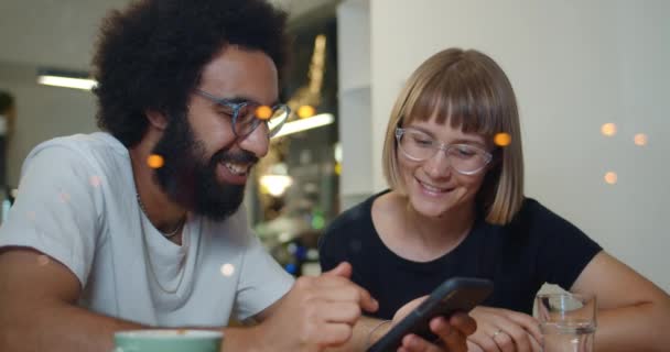 Two excited friends having great time together while sitting at modern cafe. Good looking man and woman talking and smiling while looking photos at smartphone. Concept of leisure. - Footage, Video