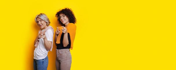 Caucasian women with curly hair are pointing at camera cheerfully while posing on a yellow free space background - Photo, Image