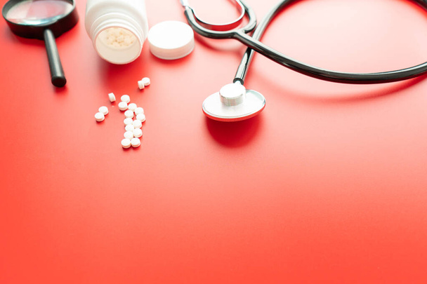 Медицинский стетоскоп и таблетки на красном фоне. Здравоохранение или болезни. Таблетки или лекарства в больнице или аптеке. Кардиологическое лечение сердца. Лекарства
 - Фото, изображение