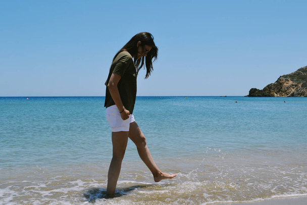 Mujer caucásica en pantalones cortos blancos y camiseta oscura de pie en una playa en un verano
 - Foto, Imagen
