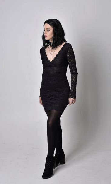 Portrait of a goth girl with dark hair wearing black lace dress and boots. Full length standing pose on a studio background. - Фото, изображение