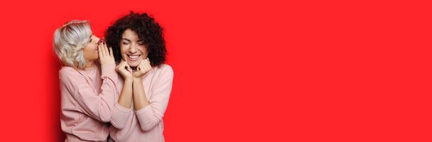 Rubia dama está susurrando algo a su rizado amigo mientras posando en una pared roja con espacio libre
 - Foto, Imagen