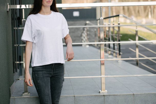 Skinny woman wearing white blank t-shirt and black jeans with space for your logo or design in casual urban style over city street background - Foto, Imagem