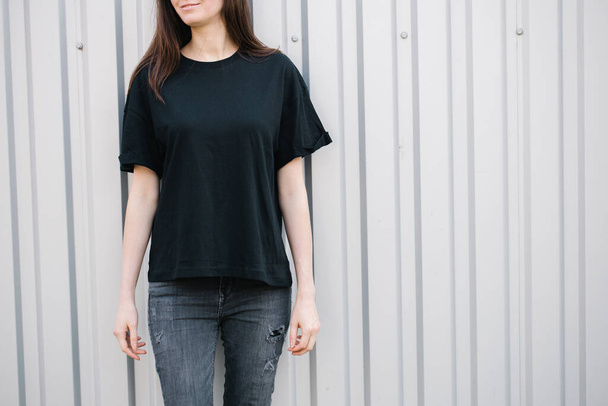 Schlanke Frau trägt schwarzes Blanko-T-Shirt und schwarze Jeans mit Platz für Ihr Logo oder Design im lässigen urbanen Stil über dem Hintergrund der Stadt - Foto, Bild