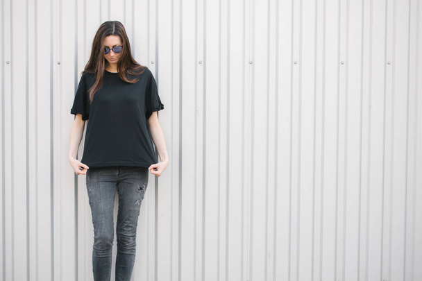 Schlanke Frau trägt schwarzes Blanko-T-Shirt und schwarze Jeans mit Platz für Ihr Logo oder Design im lässigen urbanen Stil über dem Hintergrund der Stadt - Foto, Bild