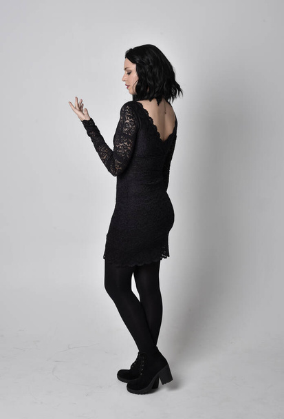 Portrait of a goth girl with dark hair wearing black lace dress and boots. Full length standing pose, facing away from the camera, on a studio background. - 写真・画像