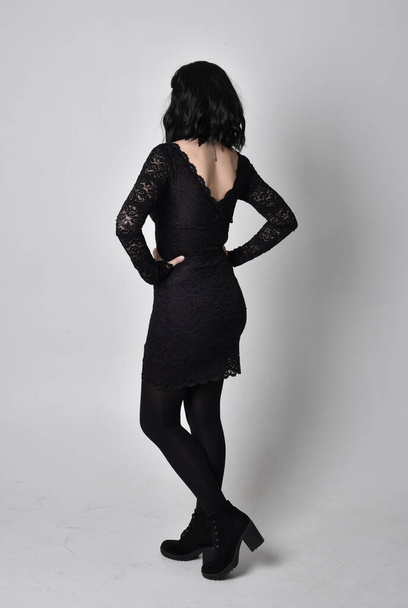Retrato de una chica gótica con el pelo oscuro con vestido de encaje negro y botas. pose de pie de longitud completa, mirando hacia fuera de la cámara, sobre un fondo de estudio
. - Foto, imagen