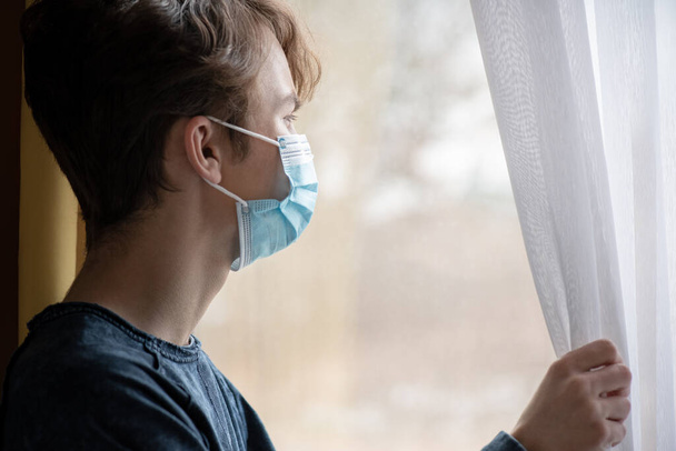 Concept of coronavirus quarantine. Child wearing medical protective face mask during flu virus, looking out of window. COVID-19 - self isolation. Teen boy forced to stay at home. - Zdjęcie, obraz