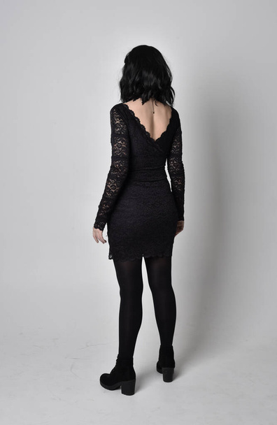 Portrait of a goth girl with dark hair wearing black lace dress and boots. Full length standing pose, facing away from the camera, on a studio background. - Photo, Image