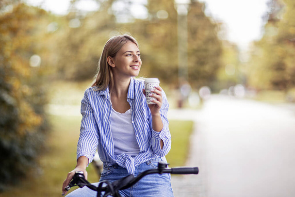 Beautiful young blonde girl on bicycle in park drinking coffee to go and smile - Фото, зображення