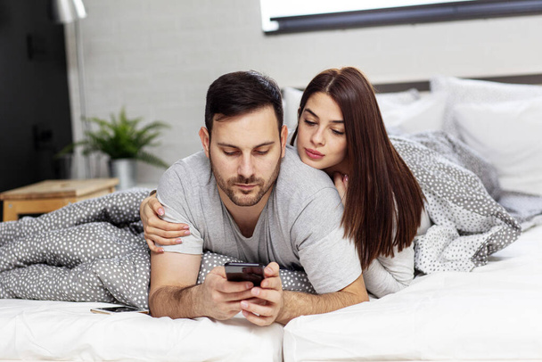 Νεαρό ζευγάρι lyuing στο κρεβάτι με κινητό τηλέφωνο στο χέρι - Φωτογραφία, εικόνα