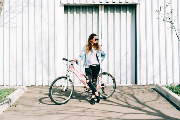 a young stylish girl holds a pink Bicycle standing next to her, against the background of a light hangar - Foto, Imagem