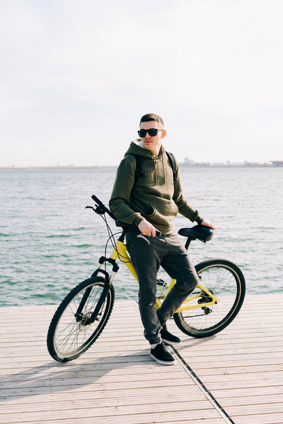 a young man in khaki sportswear and glasses stands on a pontoon near the lake and holds a yellow Bicycle near him - Foto, Imagem