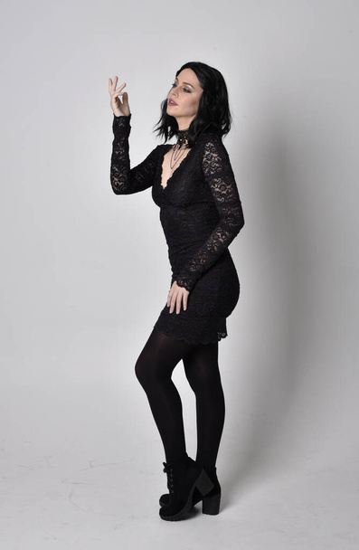 Portrait of a goth girl with dark hair wearing black lace dress and boots. Full length standing pose in profile, on a studio background. - Fotó, kép