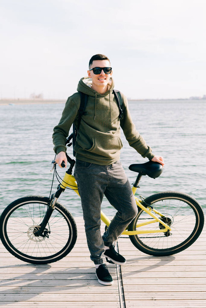 a young man in khaki sportswear and glasses stands on a pontoon near the lake and holds a yellow Bicycle near him - Photo, Image
