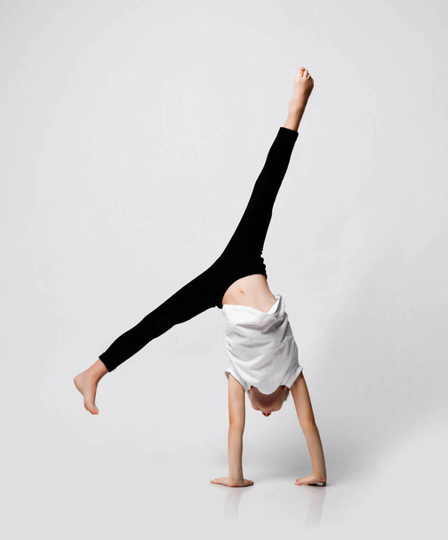 Athletic kid girl is doing gymnastic acrobatics exercises at home in studio, handstand and stretching with her legs up - 写真・画像