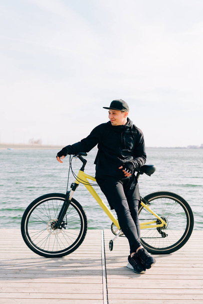 a young man in black sportswear stands on a pontoon near the lake and holds a yellow Bicycle near him - Фото, изображение