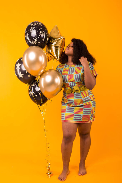 Holidays, birthday party and fun concept - Portrait of smiling young African-American young woman looking sweet on yellow background holding balloons. - Foto, Bild