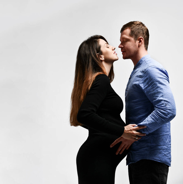Pregnant girl in black dress and her husband. He hugging her belly, they going to kiss, posing isolated on white. Close up - Valokuva, kuva