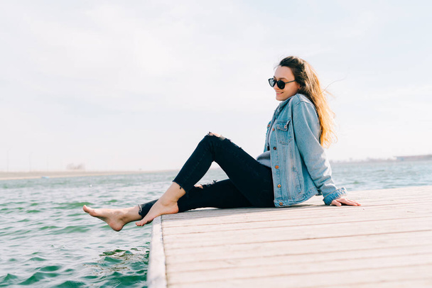 eine junge schöne stilvolle Mädchen sitzt auf einem Steg in der Nähe des Sees - Foto, Bild