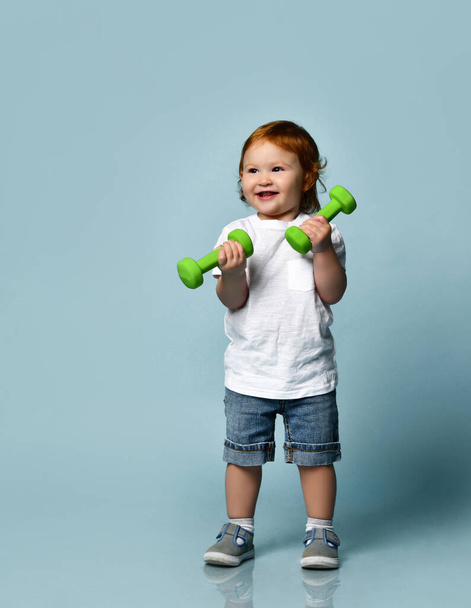 Ginger baby boy or girl in white t-shirt, shoes, denim shorts. Smiling, holding two green dumbbells, posing on blue background - Fotoğraf, Görsel