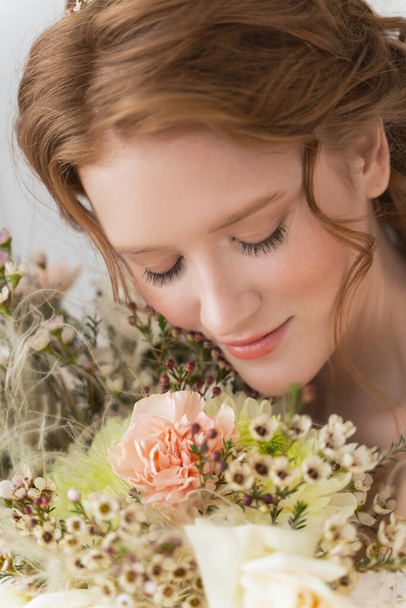 Beautiful natural redhead girl bride, with nude makeup, wearing a white dress, tenderly holds a wedding bouquet in her hands. Close up portrait - Photo, Image