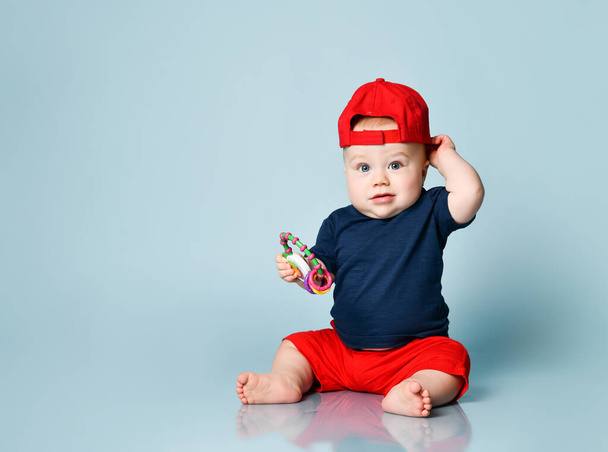 Little kid in t-shirt, red shorts and cap, barefoot. He holding a rattle, sitting on floor against blue background. Close up - Zdjęcie, obraz