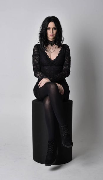 Portrait of a goth girl with dark hair wearing black lace dress and boots. Full length sitting pose  on a studio background. - Fotó, kép