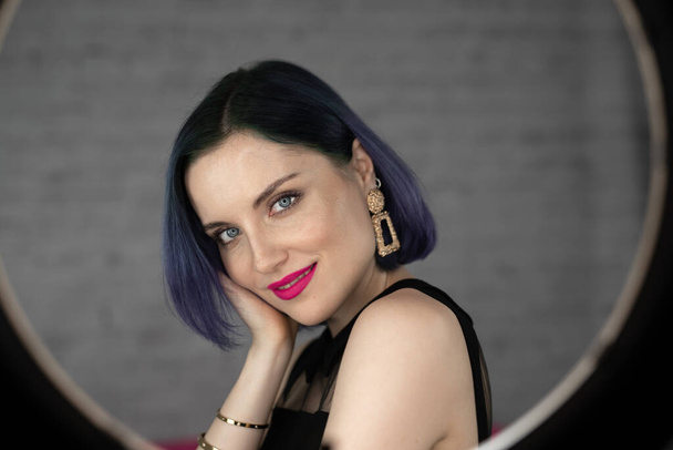 Young beautiful girl with professional makeup, blue hair and perfect skin sits in studio in front of ring light lamp posing for social media. Beauty blog. Copy space. - Fotografie, Obrázek