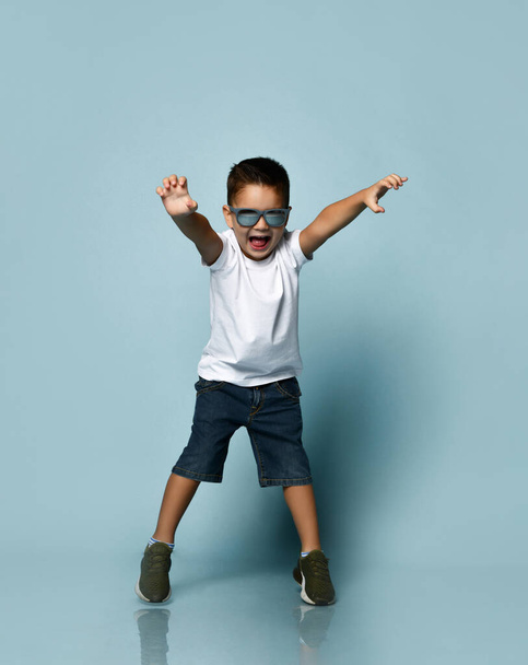 Little kid in sunglasses, white t-shirt, denim shorts and khaki sneakers. Scaring you, raised his hands, posing on blue background - Fotó, kép